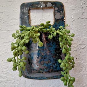 May include: A rectangular ceramic wall planter with a glossy, mottled blue and dark brown glaze. The planter features a top cutout for hanging and contains a trailing artificial plant with small, round, light green leaves cascading down.