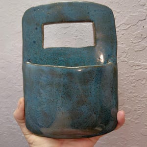 May include: A rectangular ceramic wall pocket with a glossy blue glaze featuring dark brown speckles. The object has a flat back, a curved front pouch, and a rectangular cutout at the top to serve as a handle or hanging point for wall mounting.