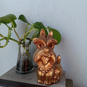 May include: A gold-colored dog figurine with a textured surface. The dog is sitting on a stack of books with gold lettering. A glass vase with a green plant is in the background.