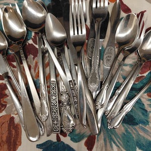 May include: A collection of assorted stainless steel flatware including various forks, spoons, and knives. The utensils feature diverse handle designs, ranging from plain polished surfaces to intricate embossed floral and geometric patterns.
