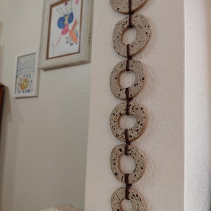 May include: A vertical wall hanging consisting of six circular, speckled off-white ceramic rings linked together by dark brown cord. The rings have an irregular, hand-formed shape with a textured surface featuring small black speckles throughout.