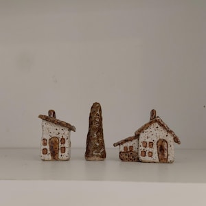 May include: Three ceramic miniature houses and a tower. The houses have white speckled walls, brown roofs, and brown doors and windows. The tower is brown and textured. The objects are displayed on a white shelf.
