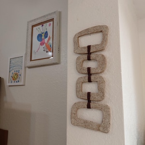May include: A vertical wall hanging consisting of four rectangular, speckled, light beige ceramic frames connected by dark brown yarn loops. The frames are arranged in a single column, each featuring rounded corners and a textured, grainy surface.