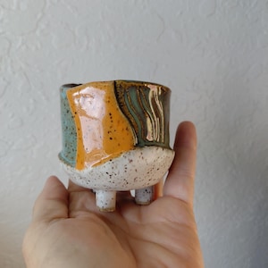 May include: A small, handcrafted ceramic planter with a speckled white base and three short legs. The upper portion features a colorful design with orange, teal, and brown glazed sections. The planter is designed for small plants or succulents.