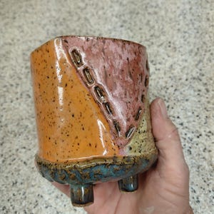 May include: A small ceramic planter with three short feet, featuring a glossy finish with a speckled orange section, a textured pink triangular panel with stitched-like indentations, and a dark blue base with a rough, organic, and uneven texture.