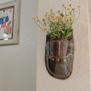 May include: A wall-mounted ceramic pocket planter with a glossy brown and teal glaze, featuring a row of small circular indentations along the front. The planter is filled with a bouquet of white daisies with yellow centers and green stems.