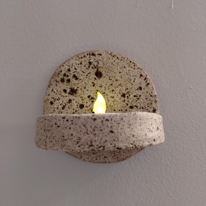 May include: A semi-circular wall-mounted ceramic sconce with a speckled cream and brown finish. The piece features a small shelf holding a glowing yellow LED tea light candle. A single hole is visible at the top center for mounting purposes.
