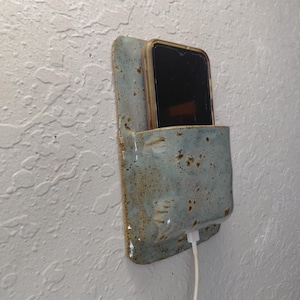 May include: A wall-mounted ceramic phone holder with a speckled light blue and brown glaze. The rectangular holder features a pocket design to secure a smartphone while charging, with a white charging cable extending from the bottom opening.