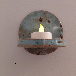 May include: A wall-mounted ceramic candle holder with a speckled light blue glaze and a brown rim. The circular backplate features a small hole for hanging, and a small horizontal shelf supports a white battery-operated tea light with a yellow flame.