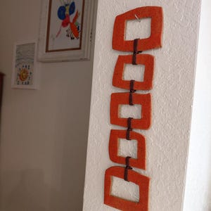 May include: A vertical wall hanging consisting of five orange ceramic square frames with a speckled texture. The frames are linked together in a chain formation by dark brown cord, creating a decorative piece suspended from a single metal nail.