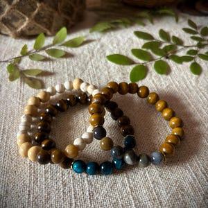May include: Three beaded bracelets stacked on a white fabric background. The top bracelet is made of white and brown beads. The middle bracelet is made of brown beads. The bottom bracelet is made of blue and brown beads.