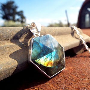 May include: A silver pendant necklace featuring a faceted, iridescent labradorite gemstone. The gemstone displays blue, green, and yellow hues. The pendant is suspended from a delicate silver chain, creating a striking contrast.