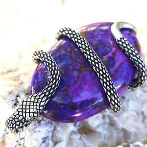 Puede incluir: Un colgante con una piedra ovalada de color púrpura con vetas oscuras, envuelta en un diseño de serpiente de metal plateado texturizado. El cuerpo de la serpiente se enrosca alrededor de la piedra y se sujeta a una cadena de metal plateado.