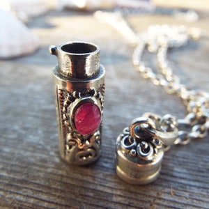 May include: A silver necklace with a small, ornate, cylindrical pendant. The pendant has a pink gemstone set into the front. The pendant is openable, and the necklace is made of a silver chain.