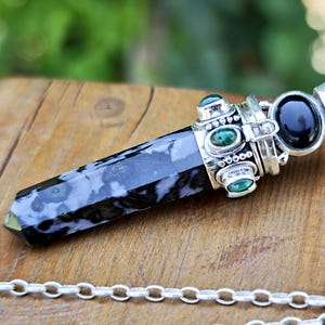May include: A pendant featuring a polished, hexagonal black and white patterned stone point set in a silver-toned metal cap. The cap is decorated with small, oval-shaped green and black cabochon stones and is attached to a silver-toned metal chain.