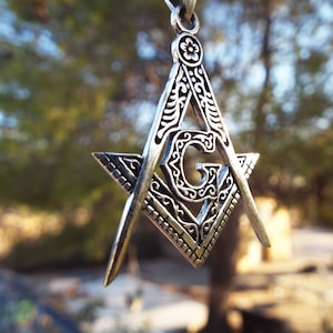 Puede incluir: Un colgante masónico de plata con detalles intrincados, que presenta un diseño de compás y escuadra con una 'G' en el centro.