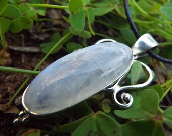 May include: A silver pendant with a large oval moonstone, set in a silver bail with a delicate swirl design. The pendant is on a black cord.