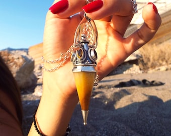May include: A silver pendant necklace with a yellow and silver pendulum-shaped charm. The charm has a silver cap with decorative stones and a pointed tip. The necklace is held up by a hand with red nail polish.
