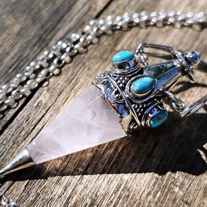 Könnte beinhalten: Ein spitzer Rosenquarz-Kristallanhänger in einer silberfarbenen Metallfassung. Die Fassung weist filigrane Metallarbeiten auf und ist mit drei ovalen Türkis-Cabochons besetzt. Der Anhänger hängt an einer silberfarbenen Gliederkette.