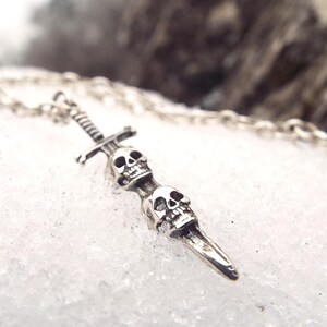 May include: Silver-toned pendant necklace featuring a dagger design with two skulls. The pendant hangs from a delicate chain, set against a snowy background. A gothic-inspired accessory.
