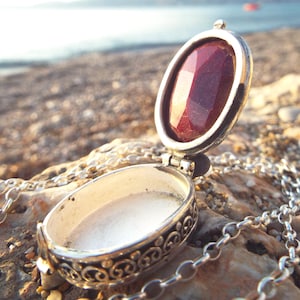 May include: A silver locket necklace with an open, oval pendant. The pendant has a faceted, deep red gemstone and is on a chain. The locket is open, showing a white interior. The necklace is displayed on a rocky surface.