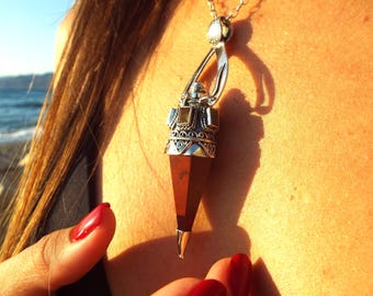 May include: A silver pendant necklace with a geometric design and a reddish-brown gemstone. The pendant hangs from a delicate silver chain. The necklace is worn against a person's skin, with the ocean visible in the background.