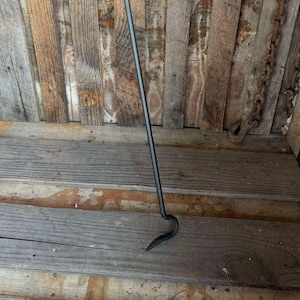 May include: A long, black metal fireplace poker with a curved hook at the end. The poker is leaning against a wooden wall.