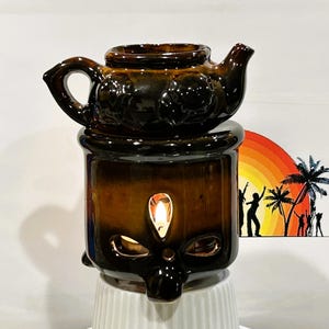 Puede incluir: Un calentador de aceite de cerámica marrón con forma de tetera con una vela en su interior. El calentador tiene un diseño floral y se asienta sobre una base con formas recortadas. Una colorida escena de playa está en el fondo.