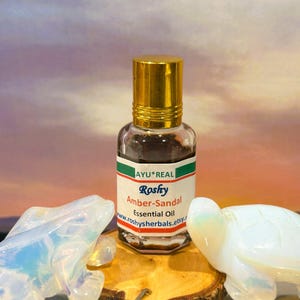 Puede incluir: Una peque&ntilde;a botella de vidrio de aceite esencial Roshy Amber-Sandal con tapa dorada. La etiqueta incluye el texto "AYU*REAL" y "Amber-Sandal Essential Oil". La botella est&aacute; sobre una superficie de madera con dos figuras decorativas de tortuga.