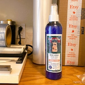 May include: A blue glass bottle with a white spray nozzle and a white label. The label has the text "Koshy's Herbals" and "BODY SPRAY ROOM SPRAY LINEN SPRAY SANDALWOOD" on it. The bottle is sitting on a wooden surface.