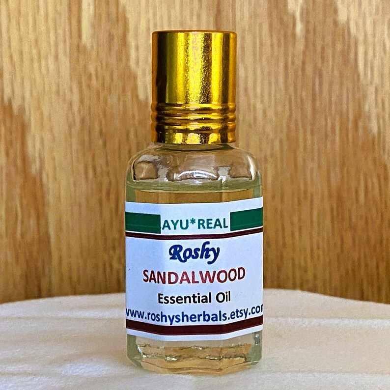 Op de afbeelding: Een klein, doorzichtig glazen flesje met een gouden dop en een groen en bruin etiket. Het etiket vermeldt "AYU*REAL Roshy SANDALWOOD Essential Oil www.roshyherbals.etsy.com".