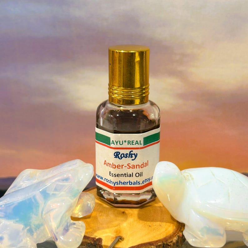 Puede incluir: Una peque&ntilde;a botella de vidrio de aceite esencial Roshy Amber-Sandal con tapa dorada. La etiqueta incluye el texto "AYU*REAL" y "Amber-Sandal Essential Oil". La botella est&aacute; sobre una superficie de madera con dos figuras decorativas de tortuga.