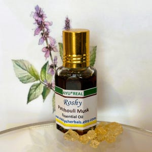 May include: A small glass bottle of Roshy Patchouli Musk essential oil with a gold cap. The label includes the text "AYU*REAL" and "roshyherbals.etsy.com". The bottle sits next to a few pieces of amber-colored resin and a floral illustration.