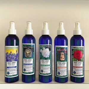 May include: Five blue glass bottles with white spray tops and labels. Each bottle contains a different scented body spray. The labels feature images of flowers, a pharaoh, a jasmine flower, a temple, and a rose. The labels also include the text "Roshy's Herbals", "Body Spray", "Room Spray", "Linen Spray", and the scent name.