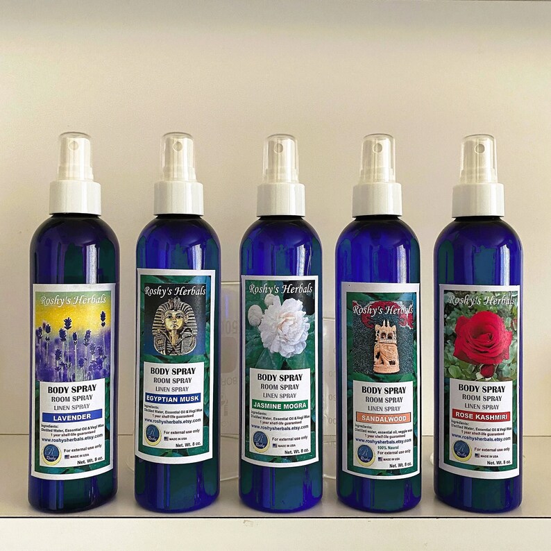 May include: Five blue glass bottles with white spray tops and labels. Each bottle contains a different scented body spray. The labels feature images of flowers, a pharaoh, a jasmine flower, a temple, and a rose. The labels also include the text "Roshy's Herbals", "Body Spray", "Room Spray", "Linen Spray", and the scent name.