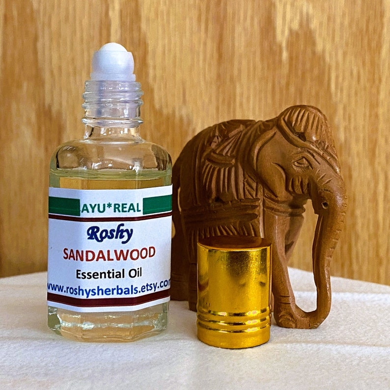 Op de afbeelding: Een heldere glazen fles met een gouden dop en een witte rolbal bovenop. De fles bevat een lichtgele vloeistof. Het etiket op de fles luidt "AYU*REAL Rosfy SANDALWOOD Essential Oil www.roshysherbals.etsy.com". Naast de fles staat een klein houten olifantbeeldje.