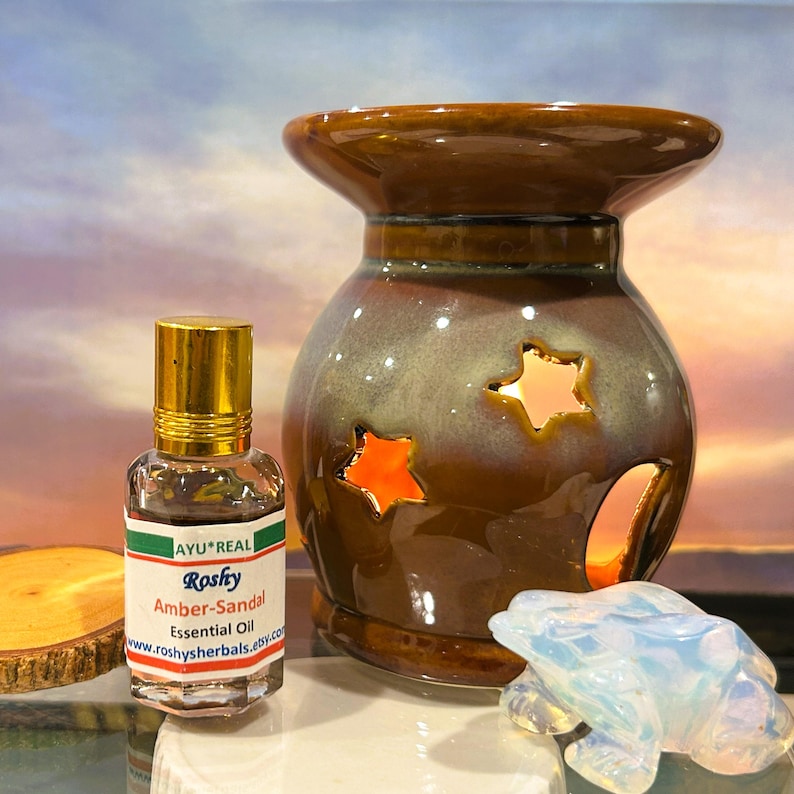 Puede incluir: Un difusor de aceites esenciales de cer&aacute;mica marr&oacute;n y gris con recortes en forma de estrella, una peque&ntilde;a botella de aceite esencial de &aacute;mbar-s&aacute;ndalo y una figura de rana transl&uacute;cida. La botella de aceite esencial tiene una tapa dorada y una etiqueta con texto.