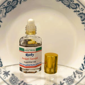 Puede incluir: Una peque&ntilde;a botella de vidrio transparente de Aceite Esencial Amber-Sandal, con un aplicador de bola blanco y una tapa dorada. La etiqueta dice "AYU*REAL Roshy Amber-Sandal Essential Oil". La botella est&aacute; sobre una superficie transparente.