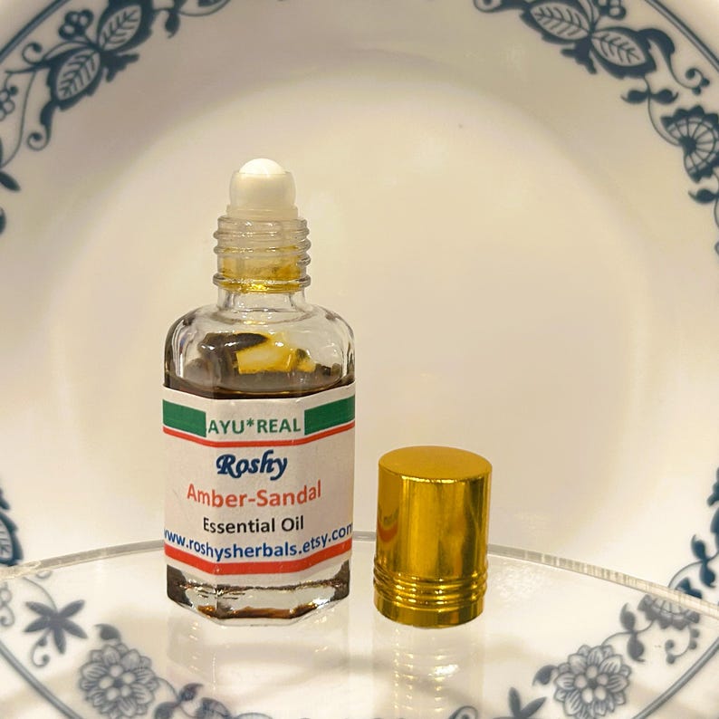 Puede incluir: Una peque&ntilde;a botella de vidrio transparente de Aceite Esencial Amber-Sandal, con un aplicador de bola blanco y una tapa dorada. La etiqueta dice "AYU*REAL Roshy Amber-Sandal Essential Oil". La botella est&aacute; sobre una superficie transparente.