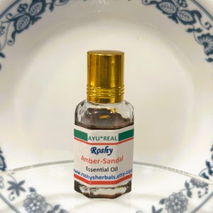 Puede incluir: Una peque&ntilde;a botella de vidrio de Aceite Esencial Amber-Sandal con tapa dorada. La etiqueta dice "AYU*REAL Roshy Amber-Sandal Essential Oil www.roshyherbals.etsy.com". El aceite en el interior es de color &aacute;mbar oscuro. La botella est&aacute; sobre un fondo blanco y azul.