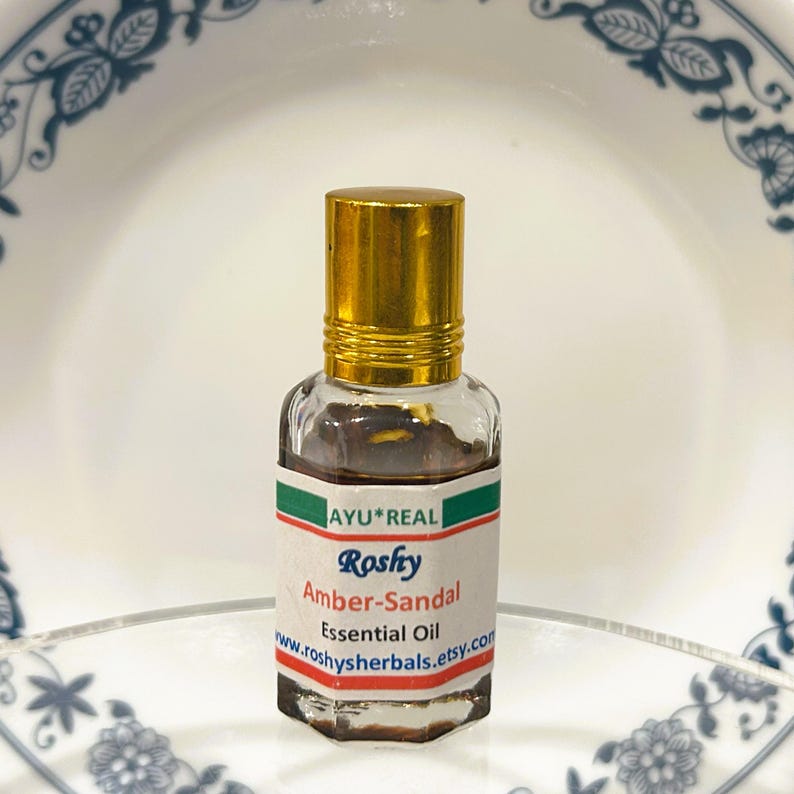 Puede incluir: Una peque&ntilde;a botella de vidrio de Aceite Esencial Amber-Sandal con tapa dorada. La etiqueta dice "AYU*REAL Roshy Amber-Sandal Essential Oil www.roshyherbals.etsy.com". El aceite en el interior es de color &aacute;mbar oscuro. La botella est&aacute; sobre un fondo blanco y azul.