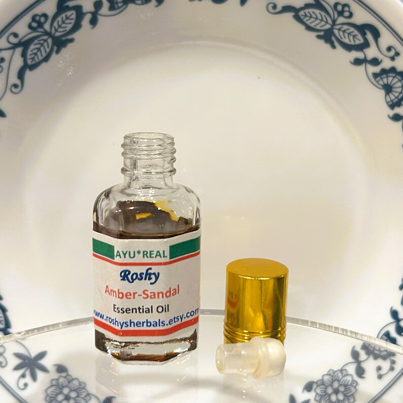 Puede incluir: Una peque&ntilde;a botella de vidrio transparente de aceite esencial Roshy Amber-Sandal con una tapa dorada y una etiqueta blanca. La etiqueta incluye el texto "AYU*REAL" y "www.roshysherbals.etsy.com". La botella est&aacute; sobre una superficie blanca.