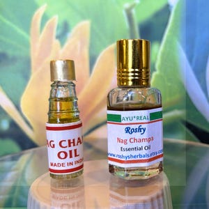 Puede incluir: Dos pequeños frascos de vidrio de aceite esencial con tapas doradas. Los frascos tienen etiquetas con el texto "Nag Champa Essential Oil" y "Made in India". El fondo es un patrón floral borroso.