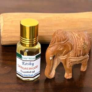 May include: A small glass bottle with a gold cap and a green label that reads "AYU*REAL Roshy SANDALWOOD Essential Oil w.roshyherbals.etsy.co". The bottle is sitting on a wooden surface next to a small wooden elephant figurine.