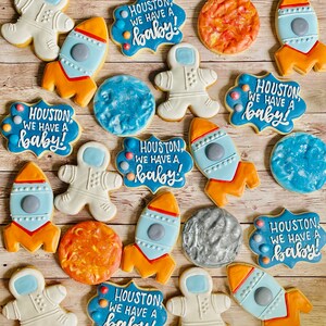 May include: A set of 14 decorated sugar cookies featuring a space theme. The cookies are decorated with blue, orange, and white icing. The cookies feature astronauts, rockets, planets, and the text "Houston, we have a baby!"