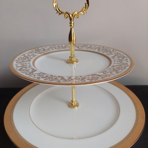 May include: Two-tiered white porcelain cake stand with a gold rim and intricate gold floral pattern. The stand has a gold metal handle.