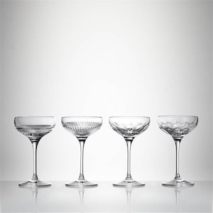 May include: Four clear champagne coupe glasses with different etched patterns. Each glass has a round bowl and a long stem. The glasses are arranged in a row against a white background.