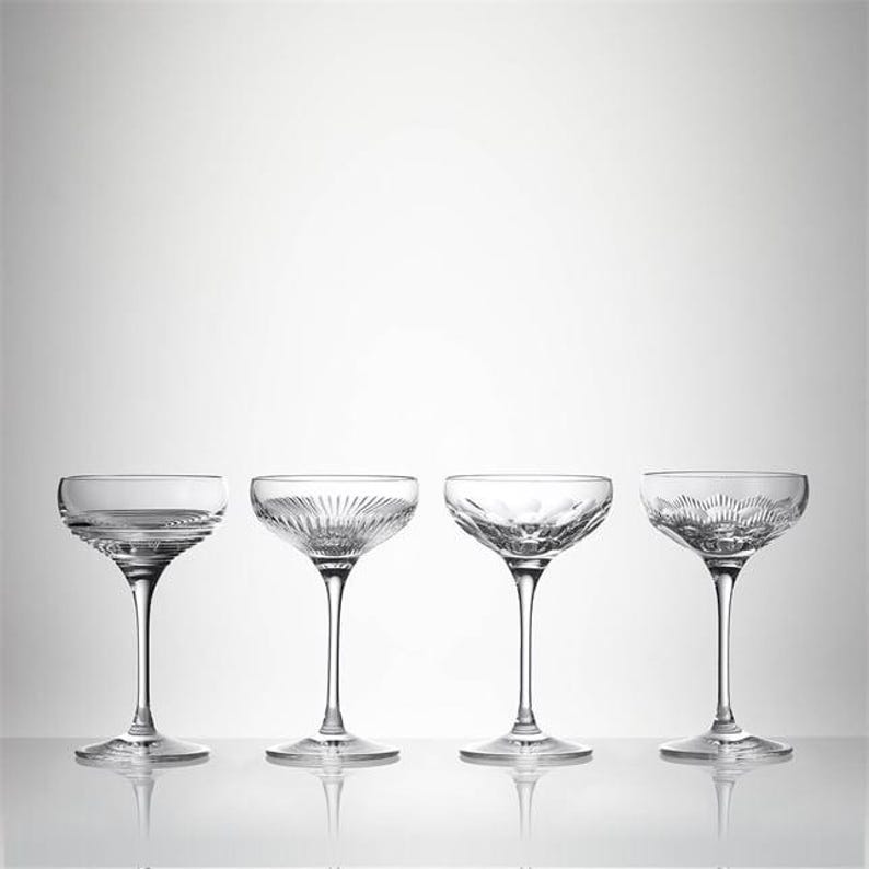 May include: Four clear champagne coupe glasses with different etched patterns. Each glass has a round bowl and a long stem. The glasses are arranged in a row against a white background.