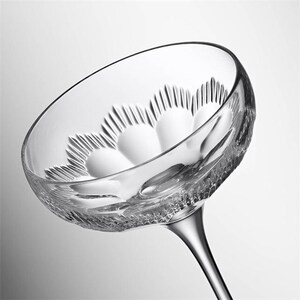 May include: A clear, stemmed champagne coupe glass with a wide, shallow bowl. The bowl features a decorative, textured pattern. The stem is slender and elegant. The glass is designed for serving champagne or other sparkling wines.