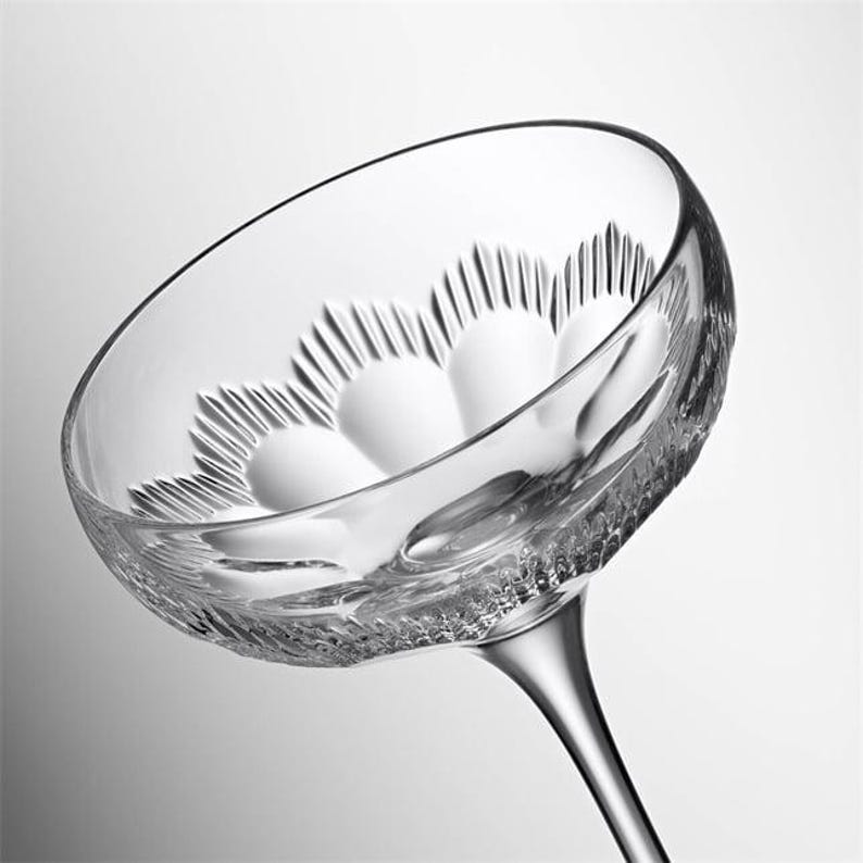 May include: A clear, stemmed champagne coupe glass with a wide, shallow bowl. The bowl features a decorative, textured pattern. The stem is slender and elegant. The glass is designed for serving champagne or other sparkling wines.